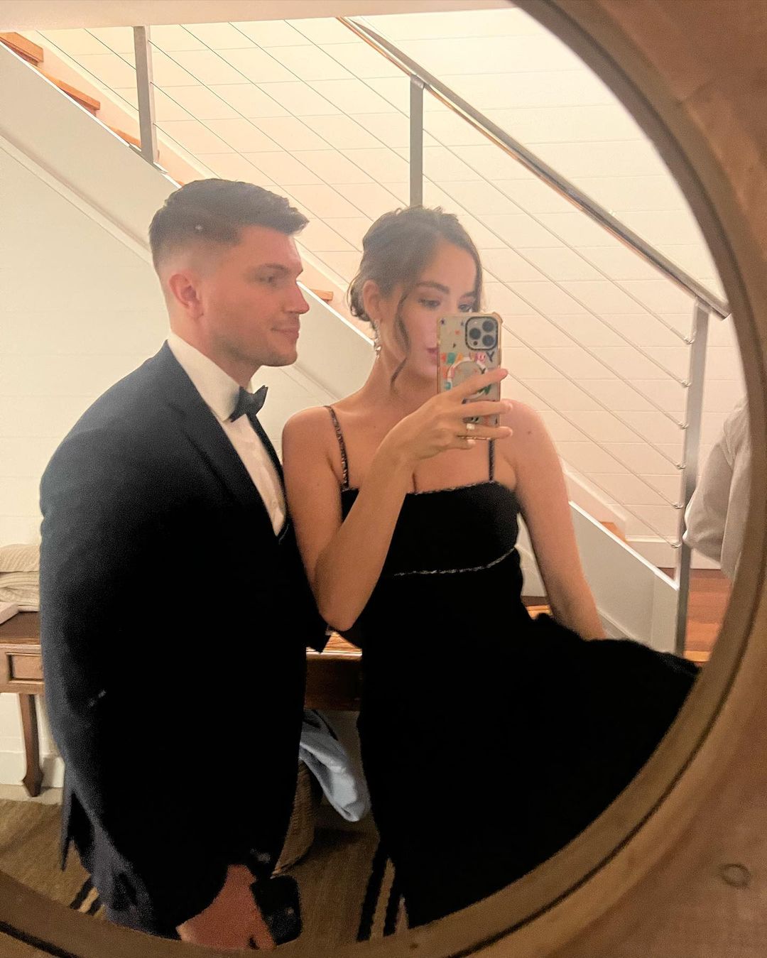 Elegant couple in formal wear poses in front of a mirror, the woman holding up a cellphone to take a selfie. Indoor setting.