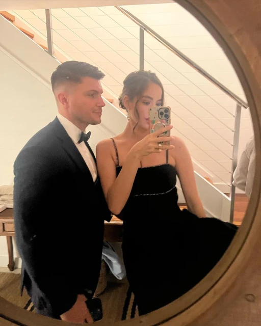 Elegant couple in formal wear poses in front of a mirror, the woman holding up a cellphone to take a selfie. Indoor setting.