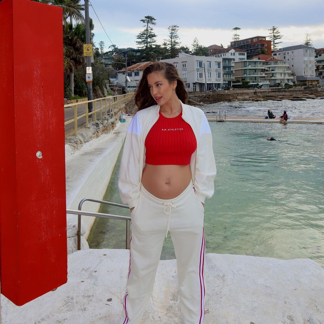 Pregnant woman in red top and white tracksuit posing near a pool in an outdoor, daytime scene with a building in the background.