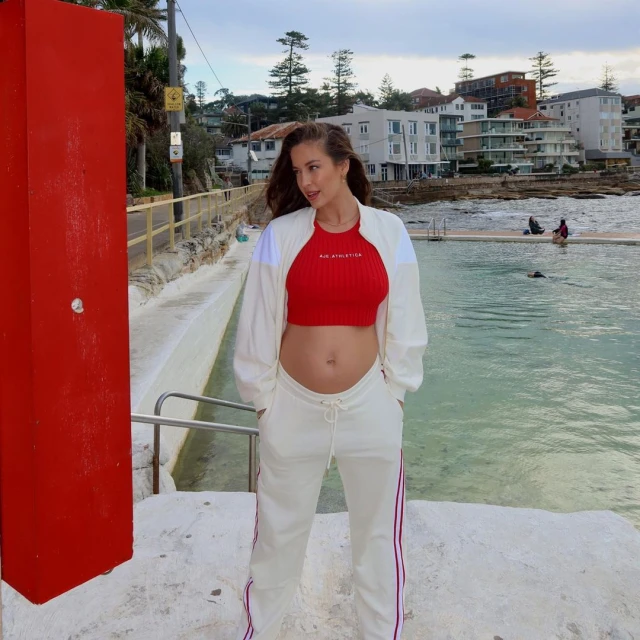 Pregnant woman in red top and white tracksuit posing near a pool in an outdoor, daytime scene with a building in the background.
