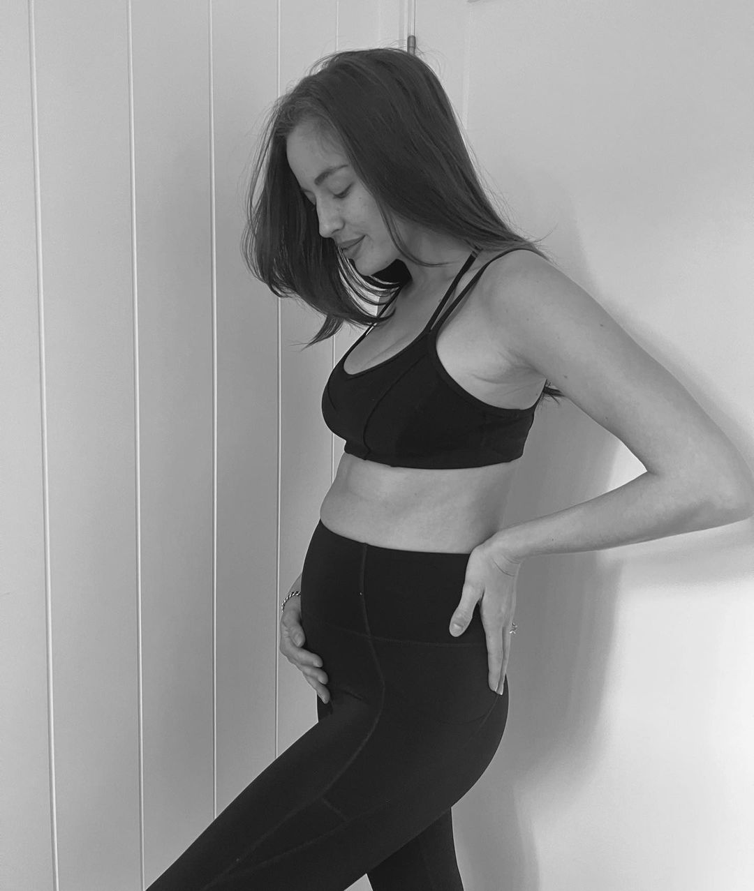 Pregnant woman in black sports bra and leggings poses indoors against a white wall, highlighting her baby bump in a black-and-white photo.