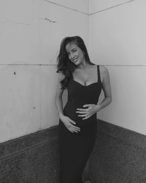 A woman in a black dress smiles and poses with her hands on her stomach, standing indoors against a textured wall in black and white.