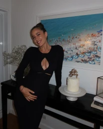 Smiling woman wearing a long black cut-out dress holding her baby bump next to a small birthday cake and large framed beach art print