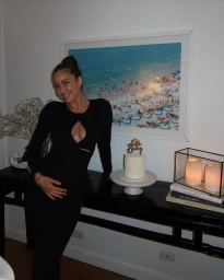 Woman in a black dress leaning against a console table with a birthday cake and framed beach photograph by Jayde Heiser