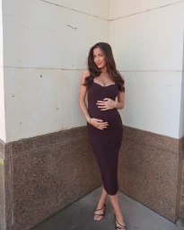 Pregnant woman in a dark brown midi dress standing against a light stone wall with hands on her belly