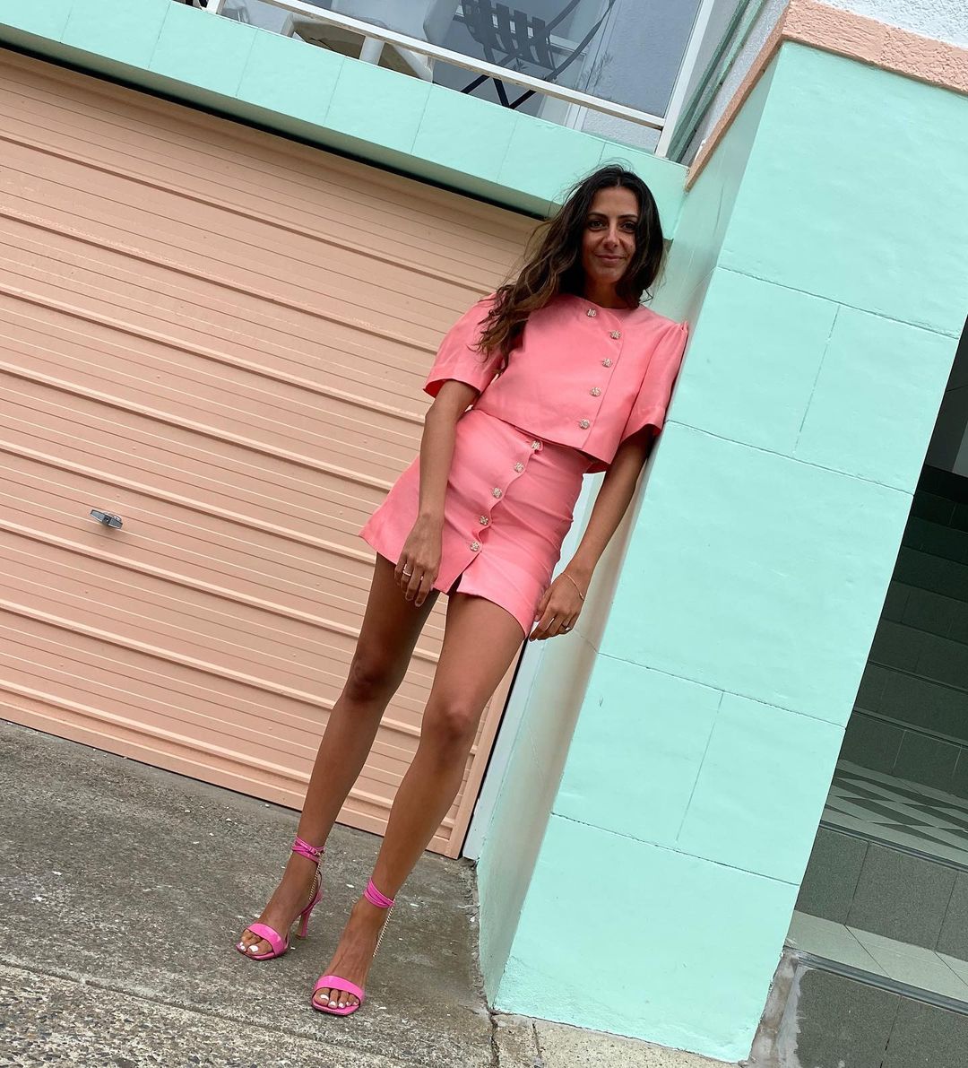 Woman in a pink skirt suit and matching heels leans against a turquoise wall in a medium outdoor shot, posing for a fashion portrait.