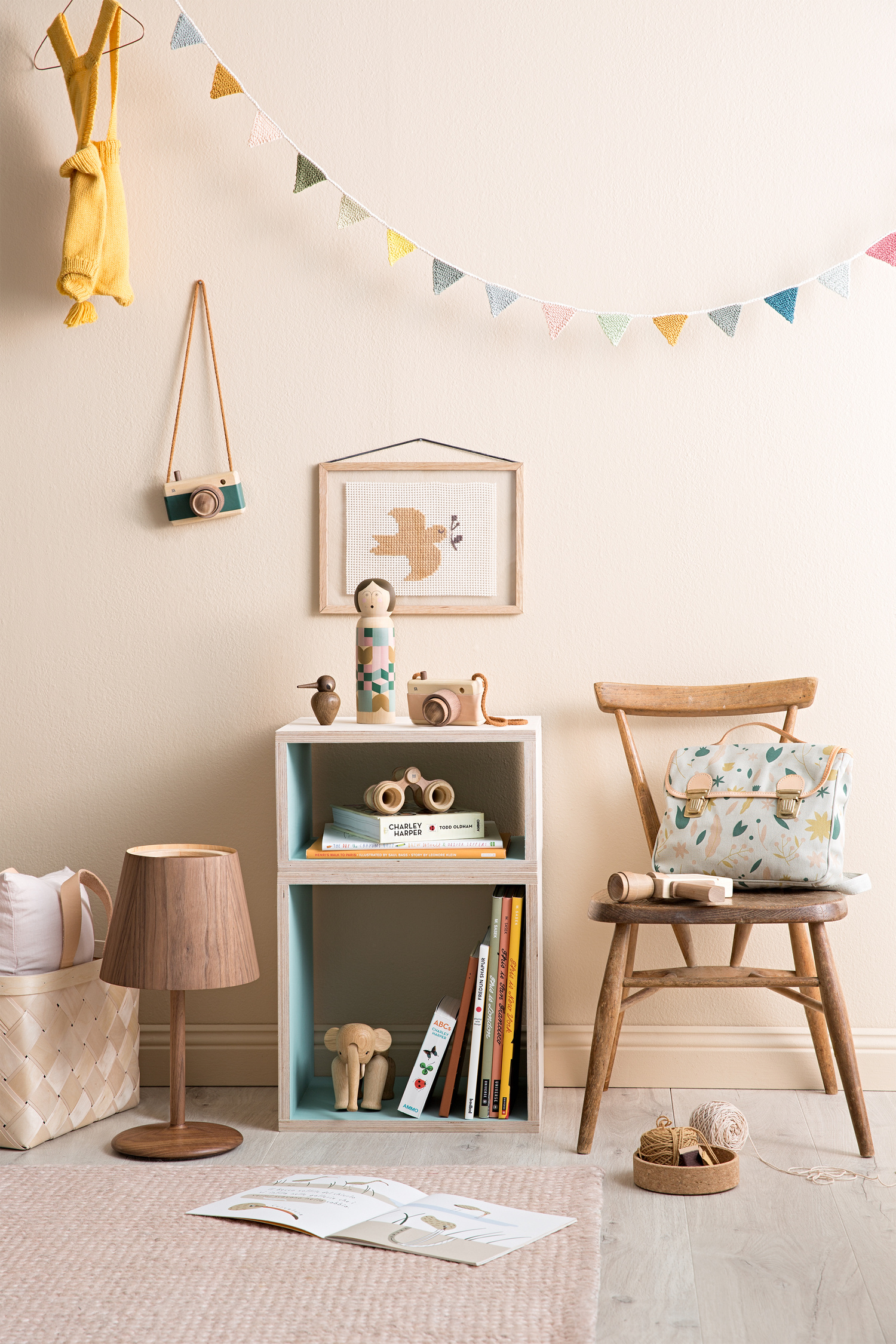 Child's room corner with wooden toys, a cube shelf with books, a wooden lamp, and colorful garland against a neutral wall.
