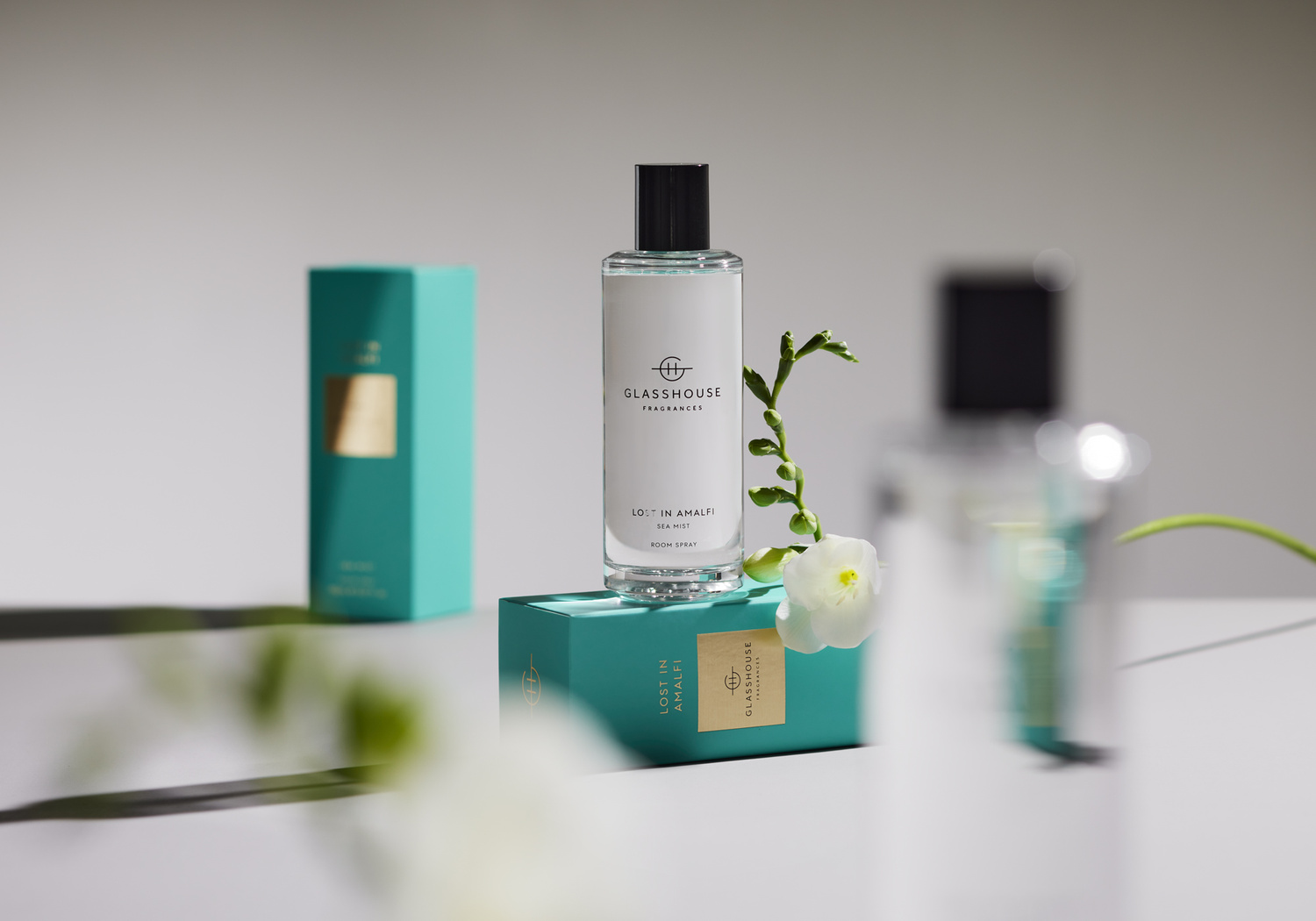 Glasshouse Fragrances 'Lost in Amalfi' sea mist room spray on a teal box, accompanied by floral sprigs and another teal box.