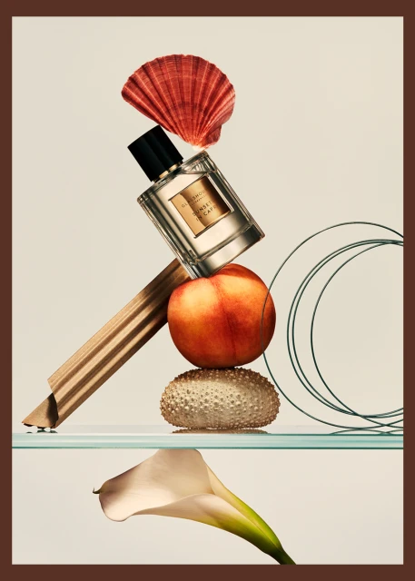 Still life composition featuring a perfume bottle, seashell, peach, sea urchin shell, and calla lily against a neutral backdrop.