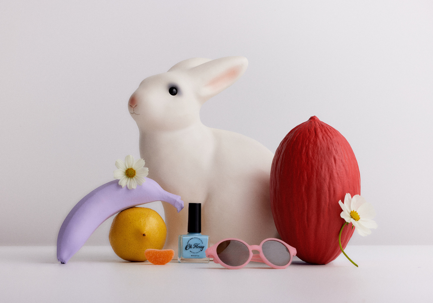 Whimsical still life of painted fruit, a bunny statue, nail polish, and sunglasses against a white backdrop, creating a surreal composition.