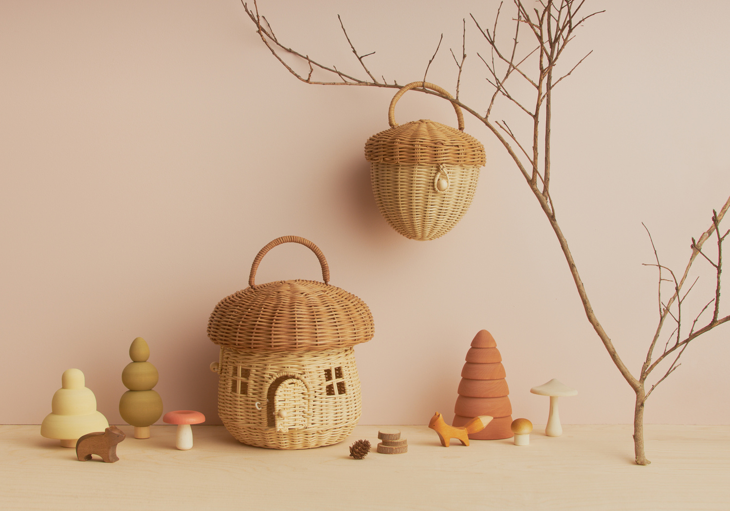 Whimsical studio shot of wicker mushroom house and acorn basket with wooden animal toys on pink backdrop with bare tree branch.
