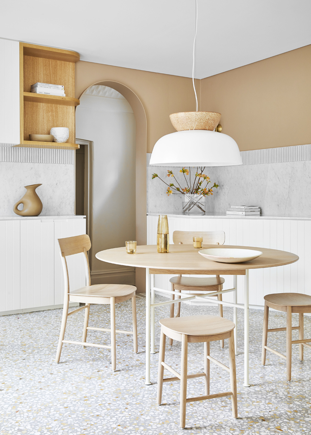 Light-toned interior scene featuring a round wooden dining table, four chairs, a stool, and neutral wall decor in a modern home.