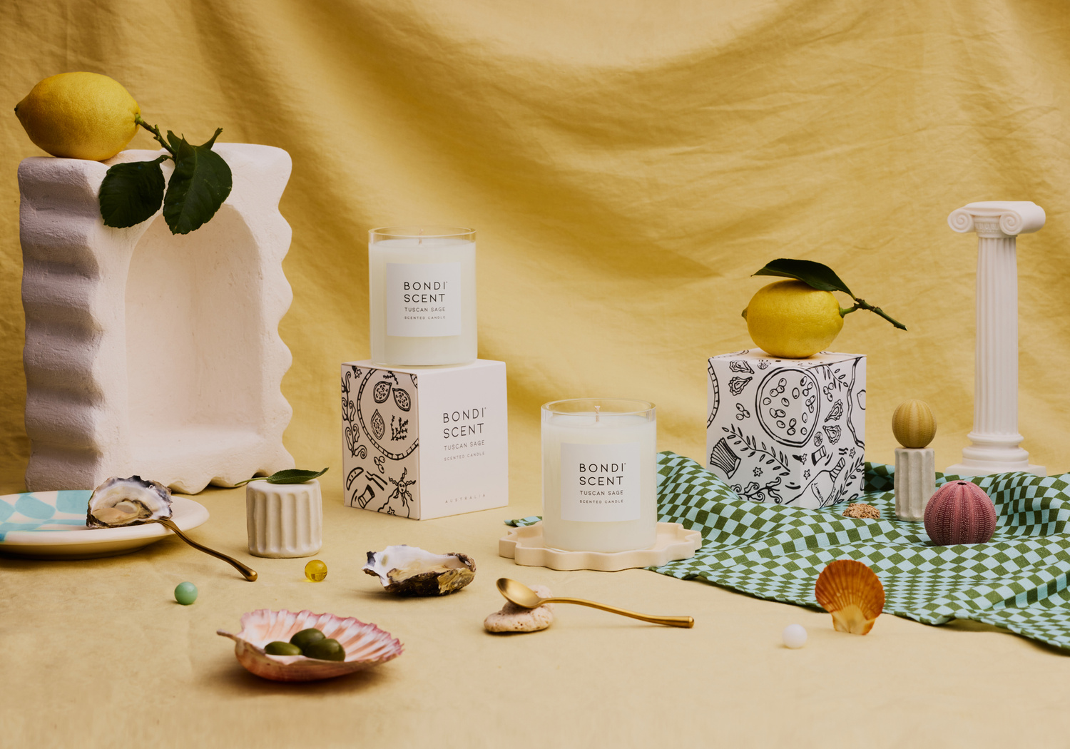 Still life featuring Bondi Scent candles, lemons, seashells, and decorative objects arranged against a draped yellow background.