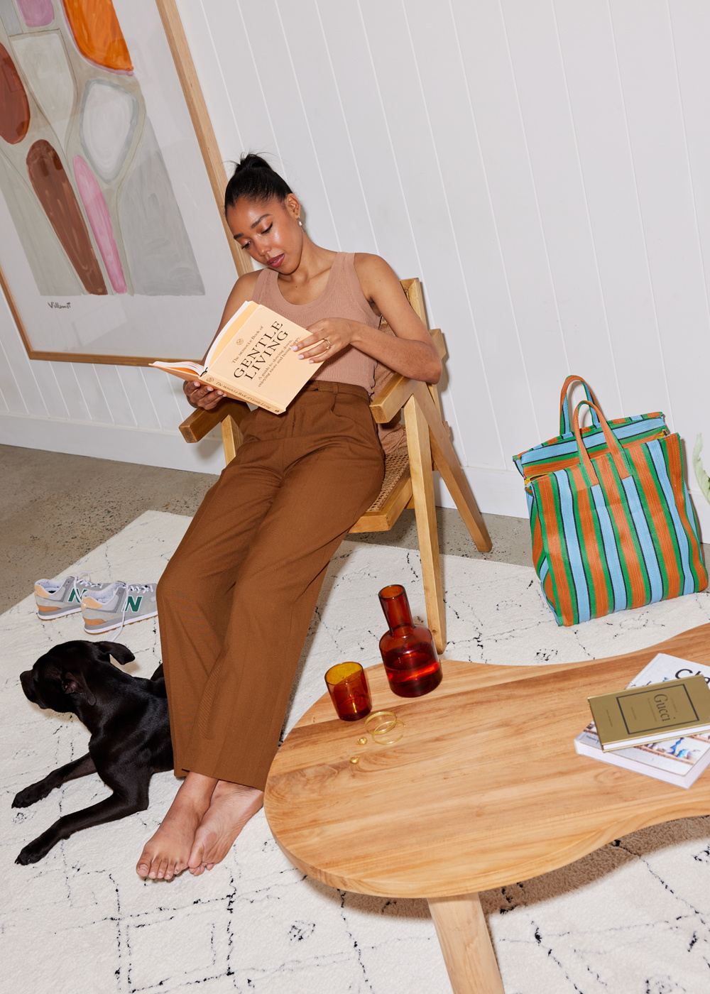 A woman reads a book while seated next to her dog in a bright living room with a white rug, wood table, and striped tote bag.