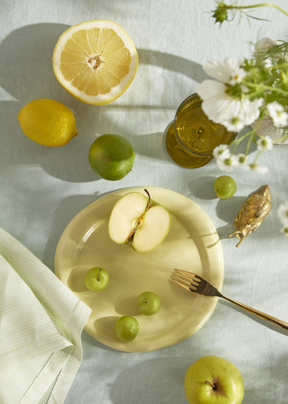 Overhead shot of a table setting with a plate of fruit, including lemons, limes, and apples, with a gold fork and decorative snail.
