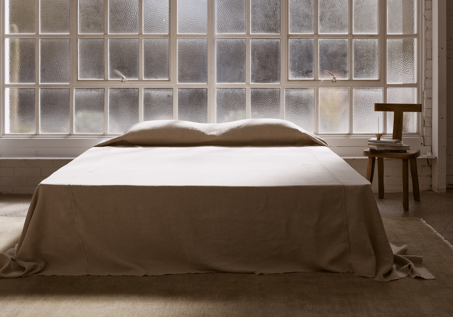 Minimalist bedroom interior featuring a large bed with beige linen, a wood stool with books, and a window with textured glass.