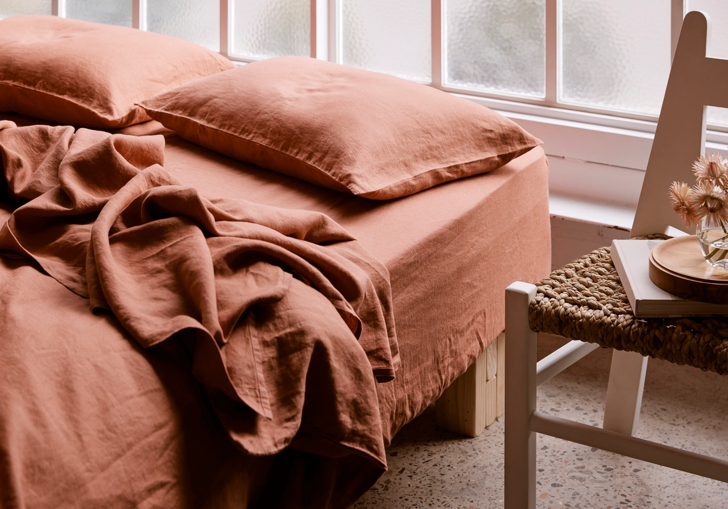 A made bed with terracotta linen sheets and pillows next to a white wooden chair with a book and dried flowers.
