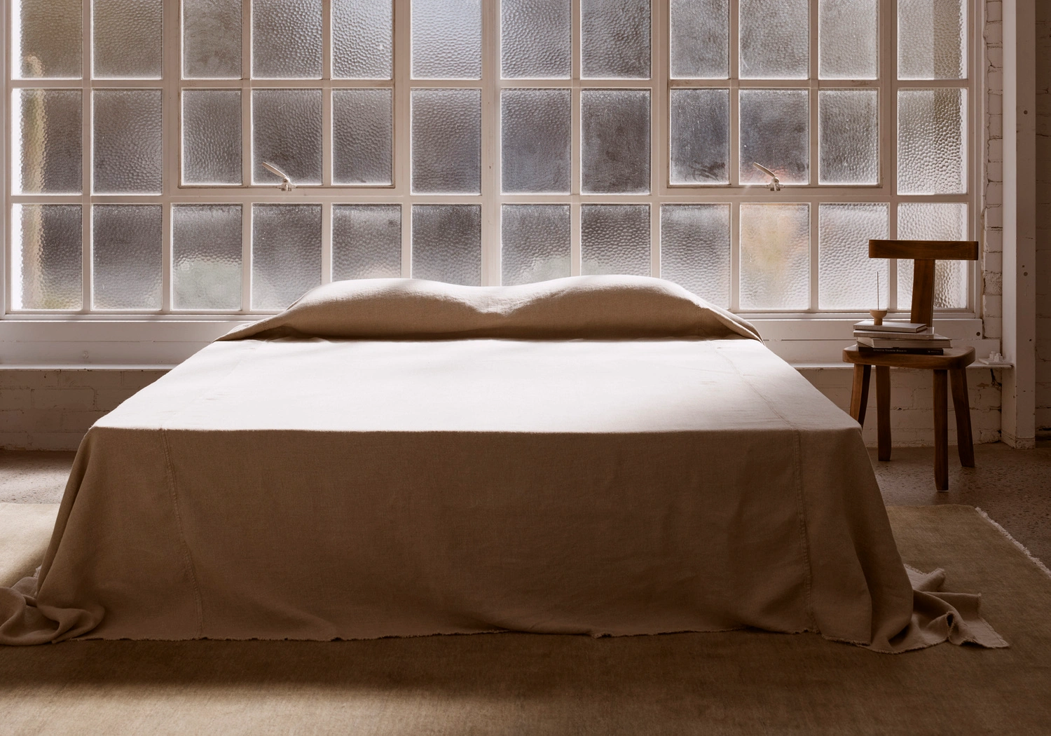 Minimalist bedroom interior with a neatly made bed against a multi-pane grid window and a wooden chair holding books