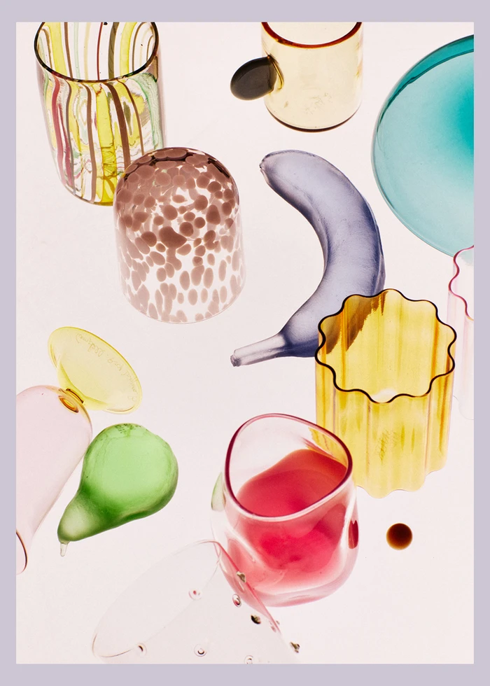 Top-down view of colourful glass tableware, tumblers, and decorative glass fruit arranged on a pale surface.
