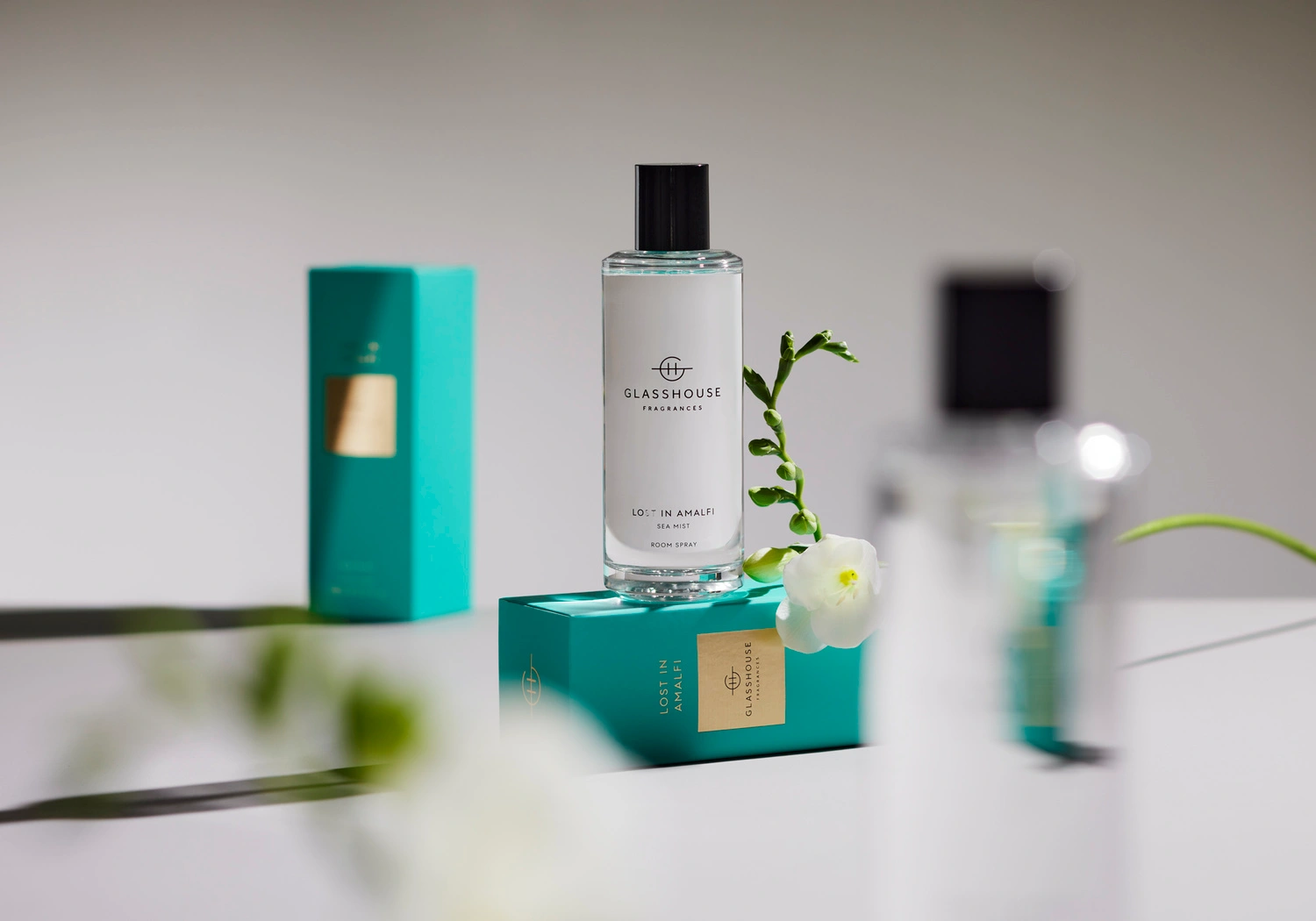 Glasshouse Fragrances room spray bottle standing on a turquoise box with white flowers against a grey background.