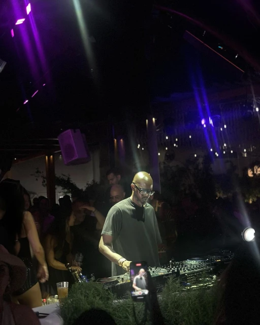 DJ performs at a lively nightclub, with vibrant lighting and a crowd of people visible in the background, creating a dynamic scene.