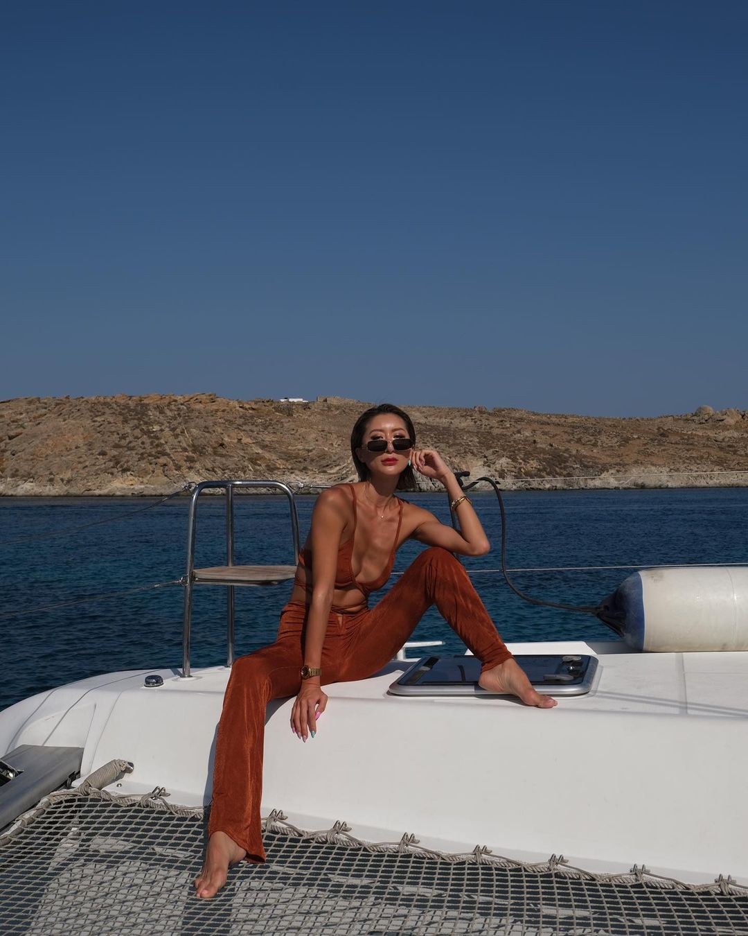 Adult woman in a brown outfit and sunglasses poses on a white boat on the sea, with a rocky landscape in the background on a sunny day.