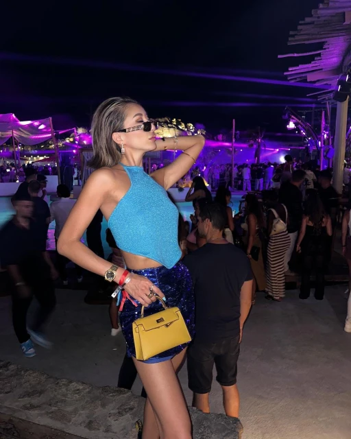 Woman in a turquoise top and blue sequin skirt poses with a yellow purse at an outdoor event at night surrounded by a crowd.