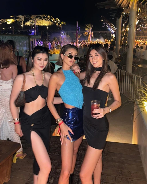 Three adult women in fashionable party attire posing together at an outdoor nightclub with a crowd visible in the background at night.