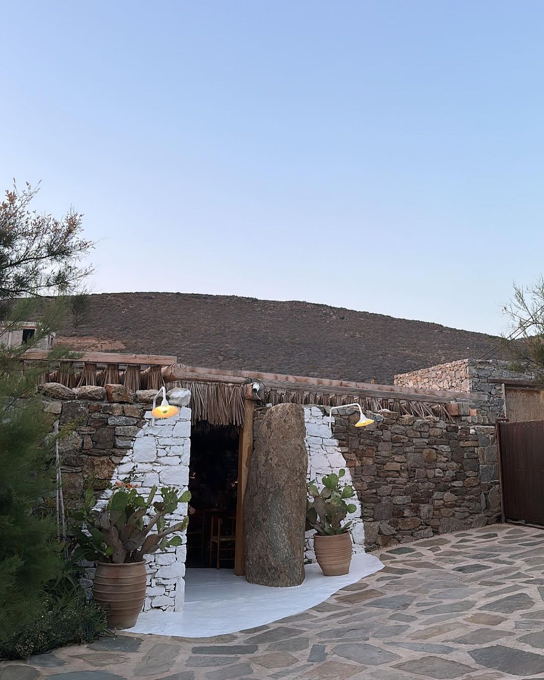 Stone building with an entrance flanked by cacti in pots, illuminated by warm lamps under the evening sky in a rustic outdoor setting.