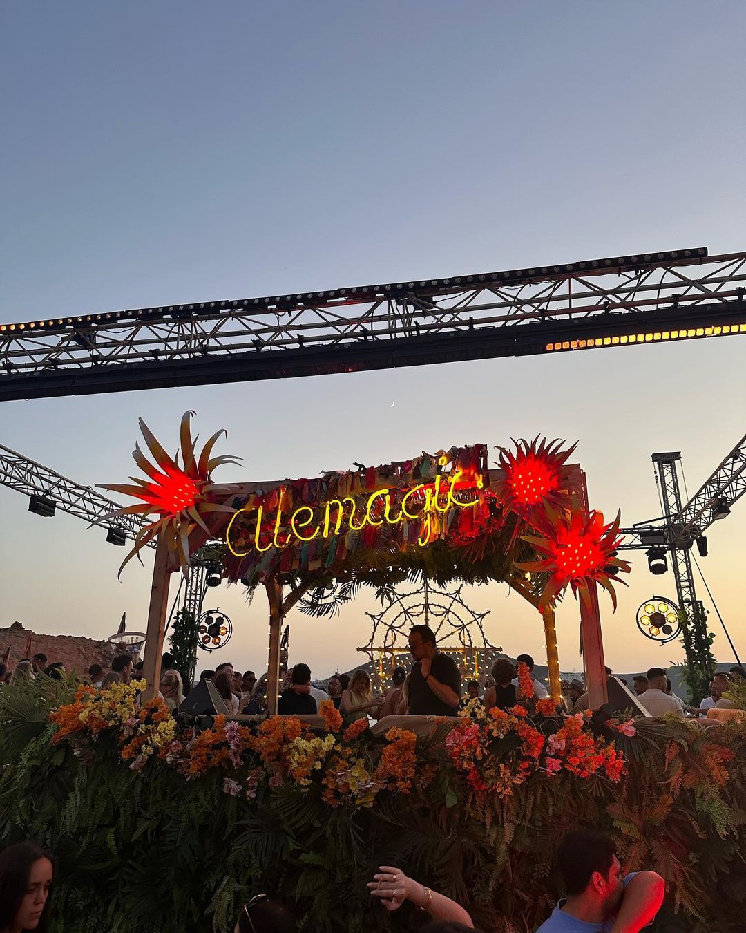 Festival stage decorated with neon sign, flowers, and lights, with crowd visible in the background against a twilight sky.