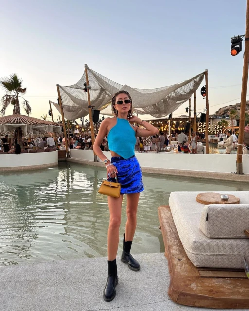Woman in blue top and sequin skirt standing at an outdoor event with a resort, swimming pool, and other people visible in background.