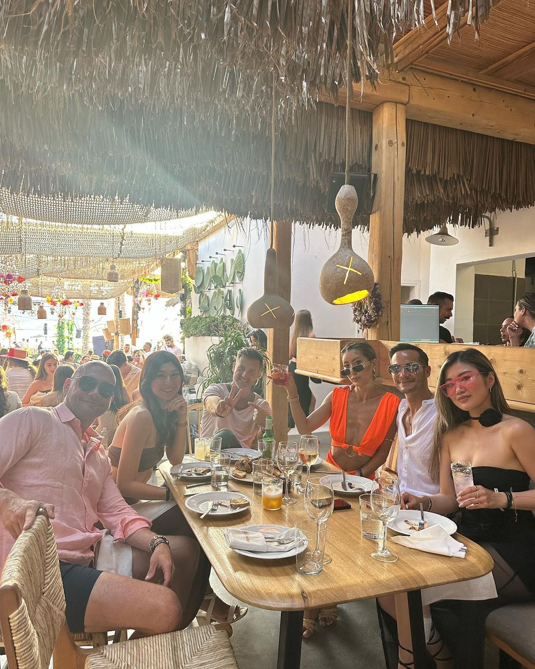 Group of six adults at outdoor restaurant. People at wooden table with drinks and plates under straw-covered canopy. Relaxed, social setting.