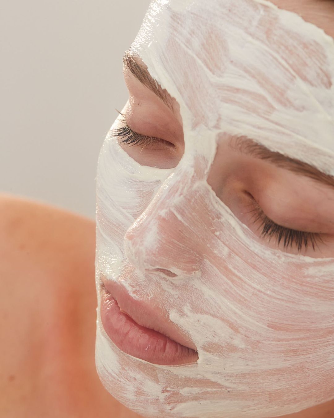 Close-up of a woman's face covered in a white beauty mask, eyes closed. The image showcases skincare and self-care in a minimalist setting.