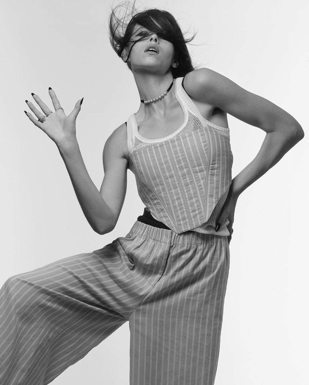 A woman poses dynamically in a striped top and pants against a white background, showcasing a blend of fashion and confident expression.