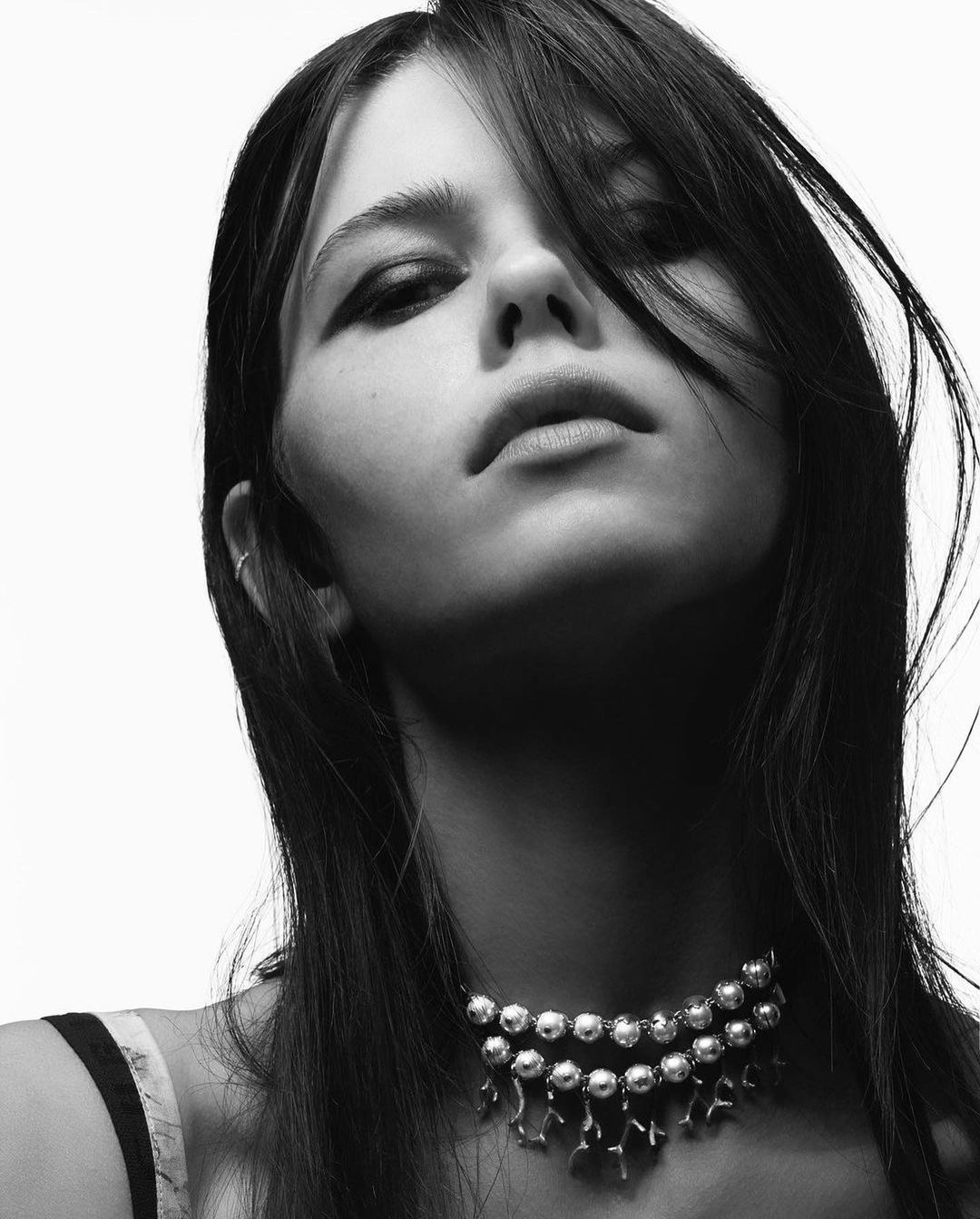 Black and white portrait of a young woman with dark hair wearing a silver necklace, shot from a low angle against a white background.