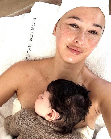 Adult woman and baby in a bright, neutral spa setting. Woman has a towel on her head and holds a baby in a warm sweater.