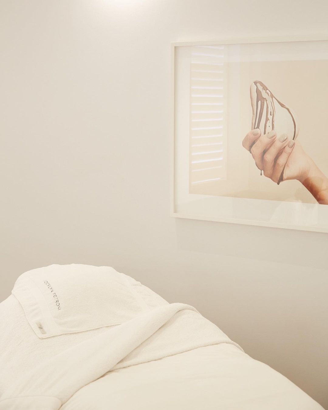 Artwork featuring a hand holding an object on a wall above a treatment table with white linens inside of a room with neutral tones.