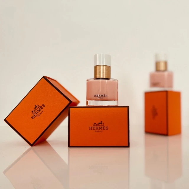 Still life shot of two Hermes Paris perfume bottles, one standing on its matching orange box, another next to a tilted box on a white surface.