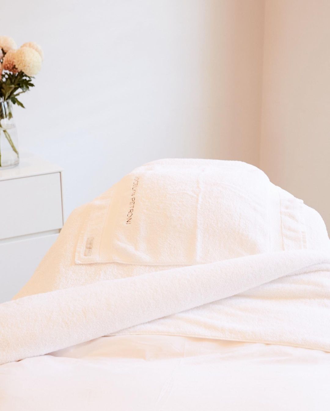 Spa treatment bed with clean white towels, indoor setting. Flowers in a vase and white cabinet visible in the background. Minimalist aesthetic.