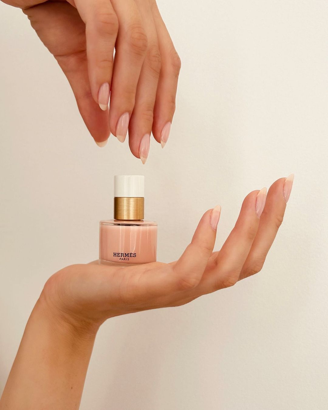 Close-up shot of two hands presenting a bottle of peach-colored Hermès nail polish on a neutral background in a beauty context.