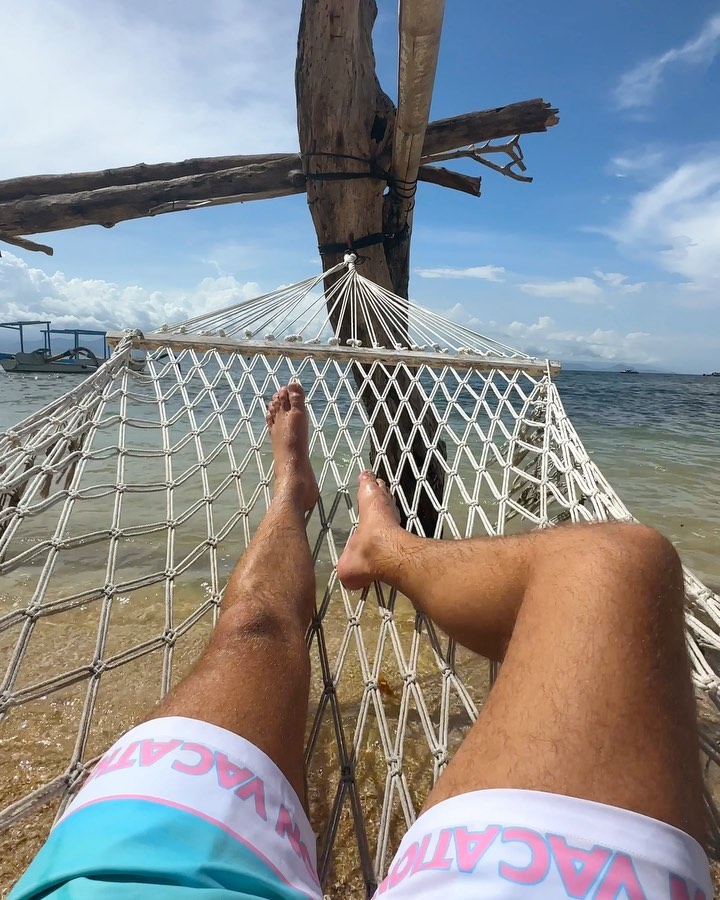 Man relaxing in a hammock on a tropical beach with turquoise ocean and blue sky. Vacation shorts are visible in the foreground.