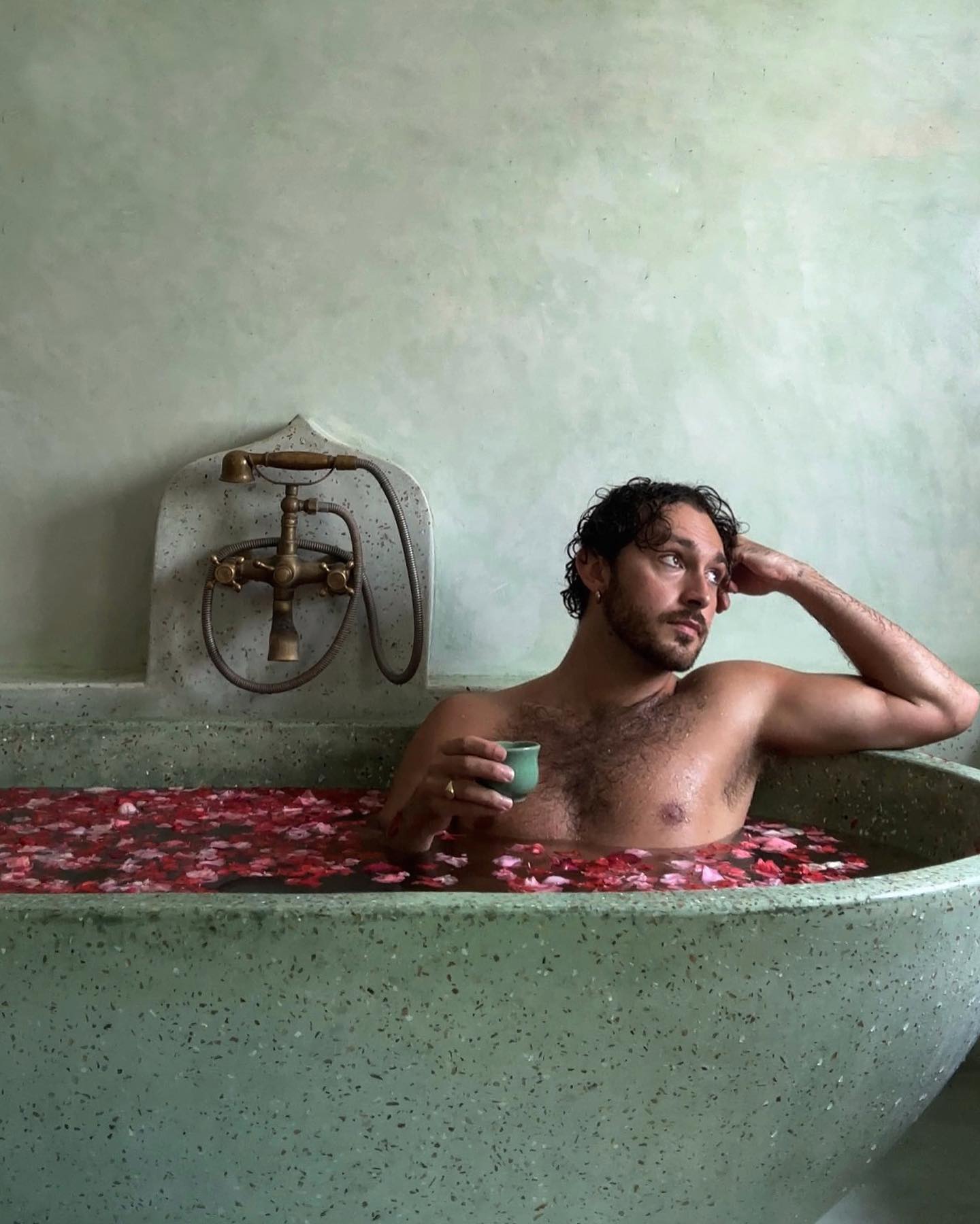 Adult male relaxes in a vintage bathtub filled with flower petals, holding a cup. The setting features light green walls and retro bath fixtures.