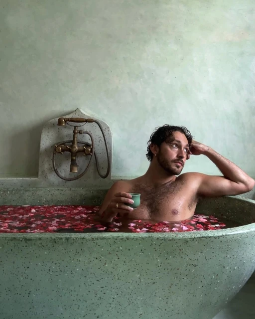 Adult male relaxes in a vintage bathtub filled with flower petals, holding a cup. The setting features light green walls and retro bath fixtures.