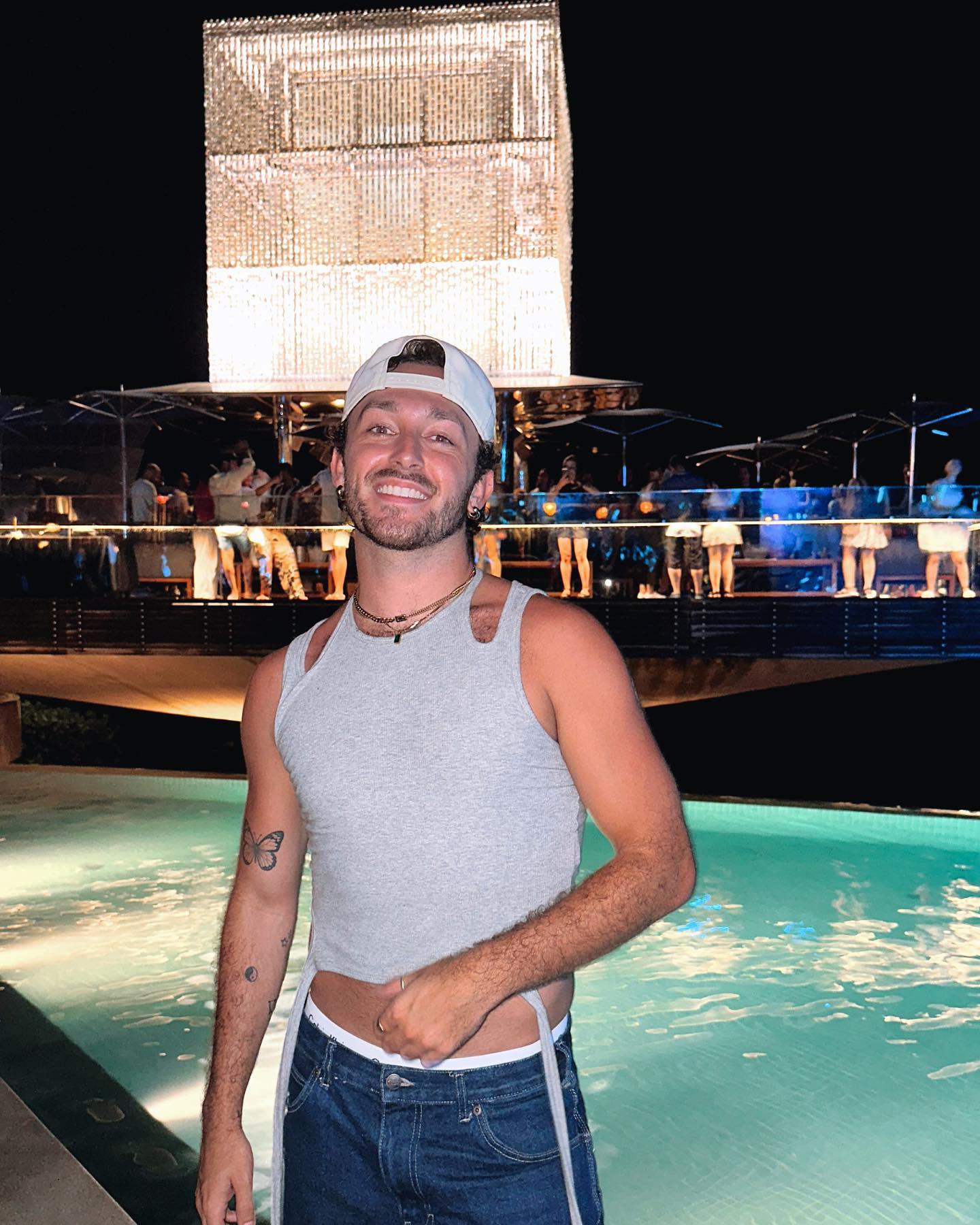 A young adult man smiles next to a swimming pool at night with a large, lit cube structure and other people in the background.