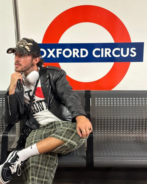 Man in a leather jacket and camouflage hat sitting in front of an Oxford Circus sign, wearing headphones, plaid shorts, and sneakers.