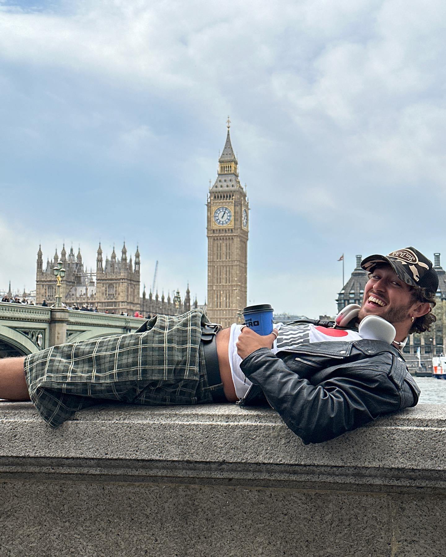 Adult male tourist in shorts lounges on embankment wall in London with takeaway coffee, Big Ben and Palace of Westminster in background.