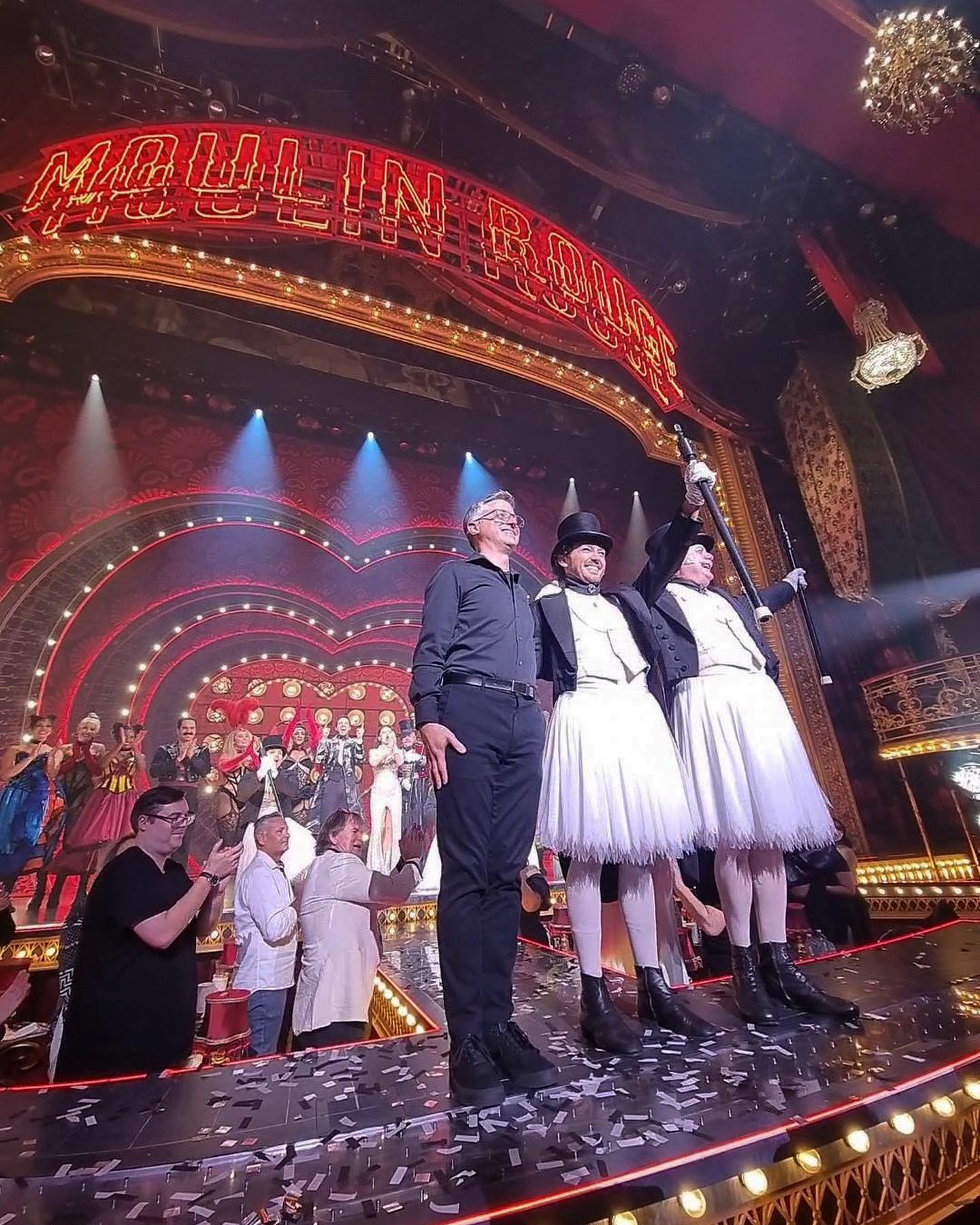 Three performers on a stage in front of a crowd with the Moulin Rouge sign in the background, theatre setting with stage lighting.