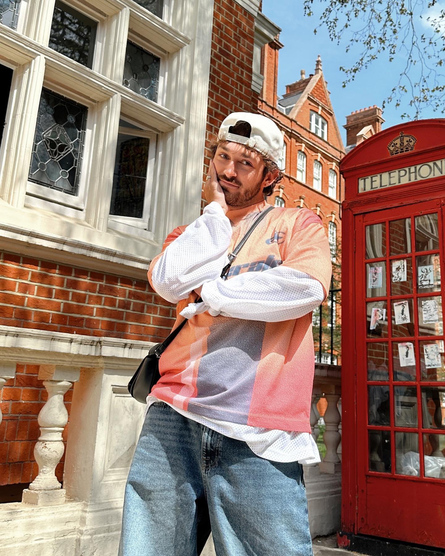 Man poses next to a red telephone booth and a brick building with large windows, wearing a white baseball cap and casual clothing.