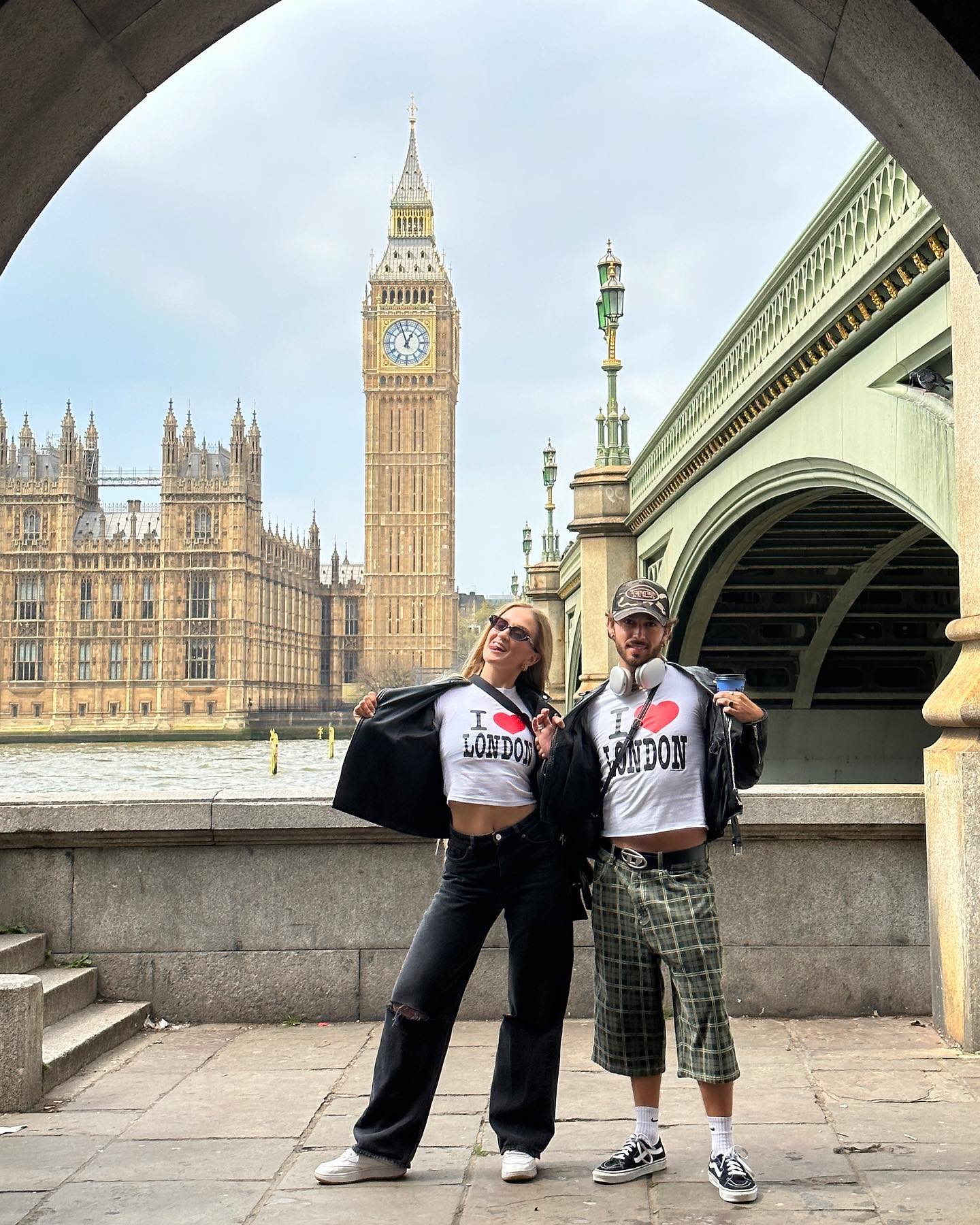 A couple in London stands near the Thames River with the Houses of Parliament and Big Ben in the background on an overcast day.