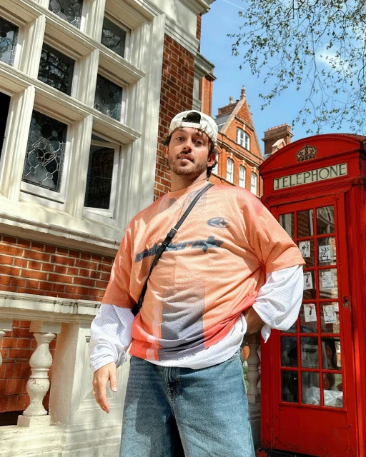 Adult male stands outside a red telephone booth on a city street wearing a baseball cap and printed shirt on a sunny day.